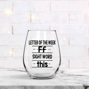 A clear stemless wine glass with black vinyl text reading Letter of the Week Ff and Sight word this, displayed on a white marble surface against a soft blurred background with warm fairy lights.