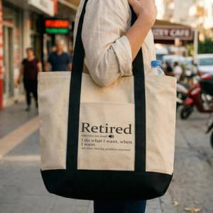 A natural canvas tote bag with black contrast handles printed with the dictionary style definition of Retired reading I do what I want when I want, carried by a person walking on a city street.