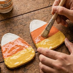 hands applying varnish with a brush on candy corn shaped clay coasters